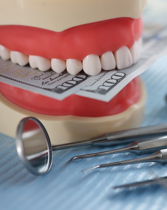 A mock jaw biting a few hundred-dollar bills next to some dental tools